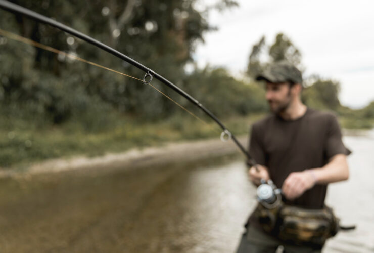 The image captures a blurred view of a man wearing a camouflage cap, focusing intently on fishing, with the fishing rod in sharp focus stretching towards the serene river in the background.